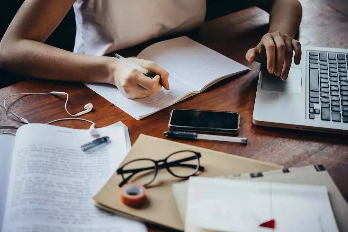 Individual writing in notebook in front of laptop.