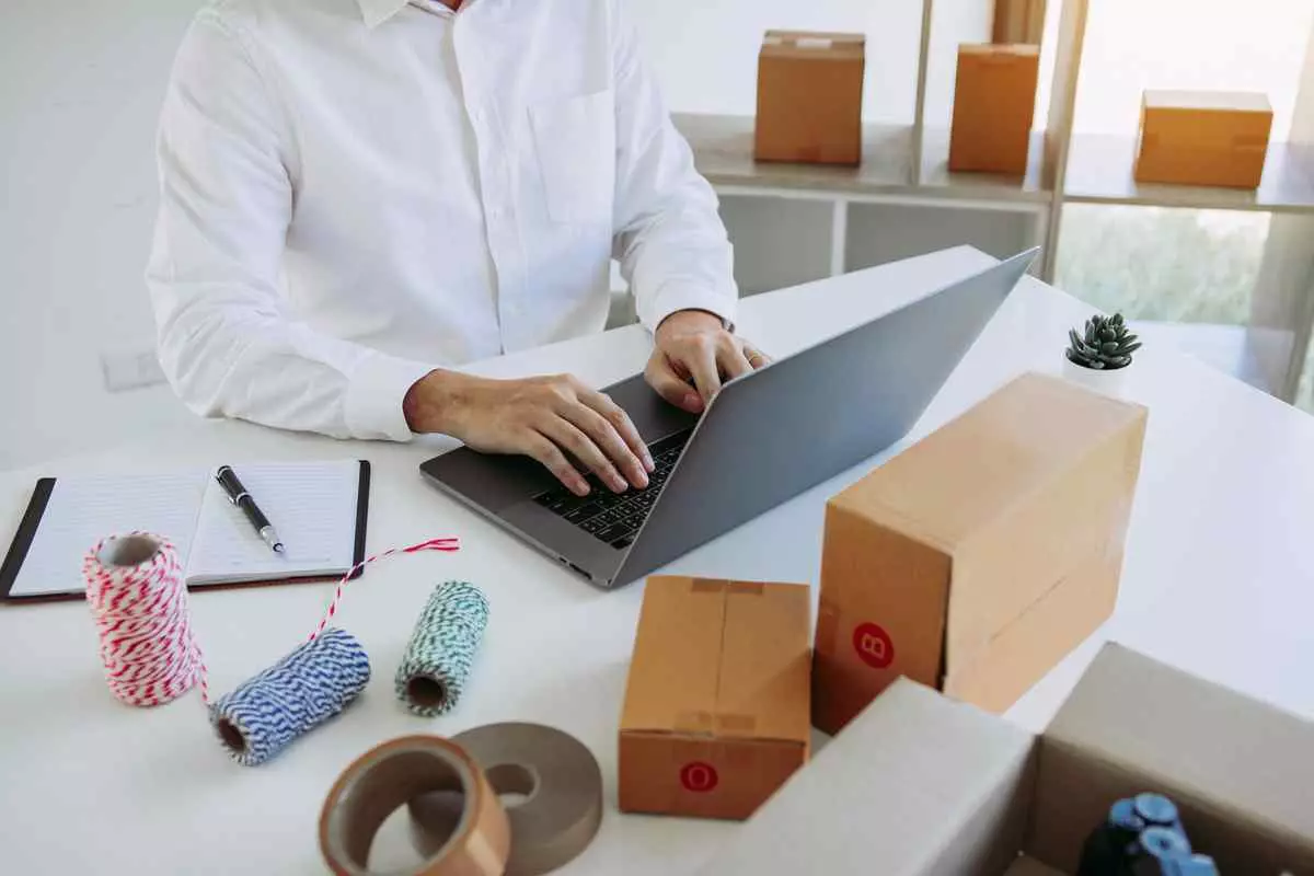 A man typing on a laptop at a desk surrounded by boxes