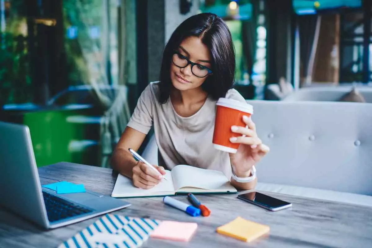 A freelancer writing notes on a notebook at a table, by a cup of coffee and a laptop